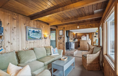 Cozy living room with wooden walls, a green sofa, a coffee table, and a view into a kitchen area.