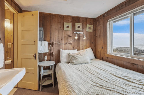 Cozy bedroom with wooden walls, a single bed, a small table, and a window showing a scenic view.