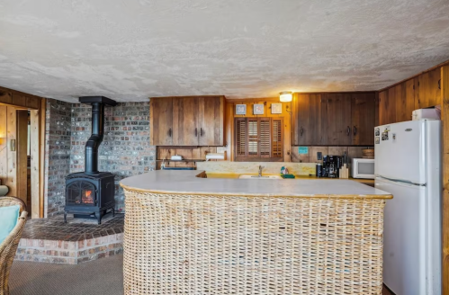 Cozy kitchen with a stone fireplace, wooden cabinets, and a woven bar counter. Warm, rustic decor throughout.
