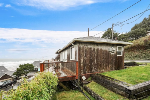 A weathered wooden house with a deck overlooks a grassy area and the ocean under a blue sky.