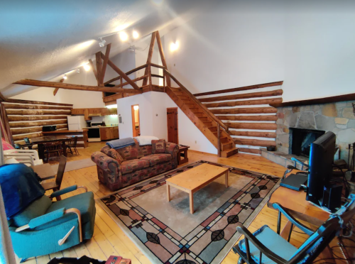 Cozy cabin interior with wooden beams, a staircase, living area, dining space, and a stone fireplace.