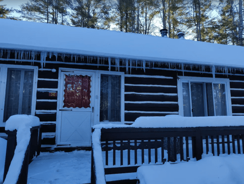A cozy log cabin covered in snow, with icicles hanging from the roof and surrounded by tall trees.