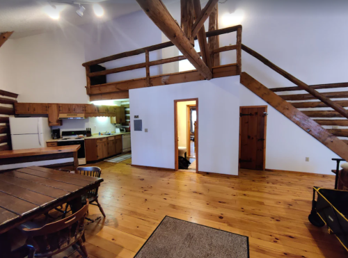 Cozy interior of a cabin with wooden beams, a kitchen, dining area, and a lofted space above.