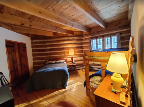 Cozy log cabin interior with a double bed, bunk beds, wooden furniture, and a warm lamp. Natural light from windows.