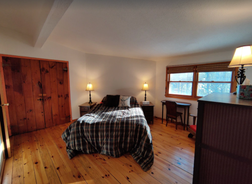 Cozy bedroom with a wooden floor, a bed with a plaid blanket, two lamps, and a desk by the window.