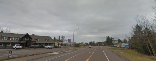 A quiet street scene with a few buildings, overcast sky, and a car driving on the road.