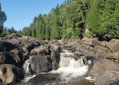 A serene river flows over rocky terrain, surrounded by lush green trees under a clear blue sky.