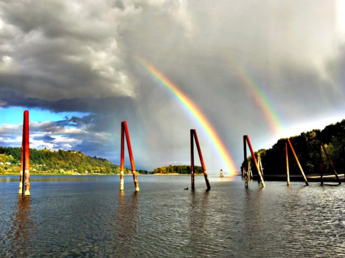 A serene river scene with two vibrant rainbows arching over weathered wooden posts under a cloudy sky.