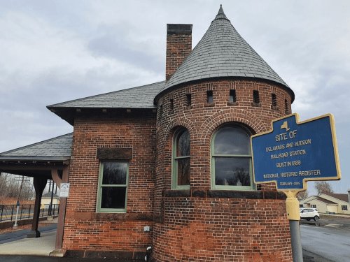 Historic brick railroad station with a conical roof and a blue sign marking its significance, under a cloudy sky.