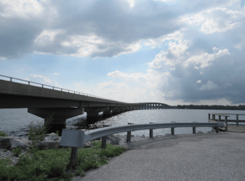 A concrete bridge spans a calm body of water under a cloudy sky, with a grassy area and guardrail in the foreground.