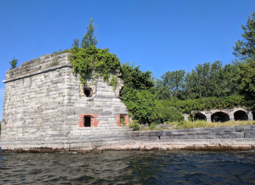 Stone ruins covered in greenery by the water, with blue sky and trees in the background.
