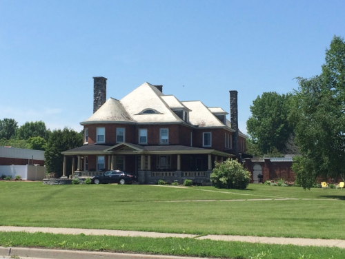 A large, historic brick house with a steep roof, surrounded by green grass and trees on a sunny day.