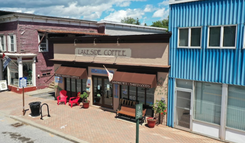 A cozy coffee shop named "Lakeside Coffee" with outdoor seating, surrounded by colorful buildings and a blue sky.