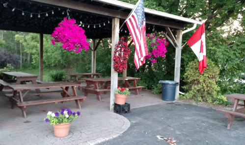 Outdoor seating area with picnic tables, hanging flowers, and flags of the USA and Canada under a wooden pavilion.