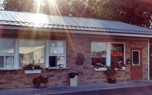 A brick building with a sign reading "Gino's Pizza," featuring flower boxes and bright sunlight reflecting off the roof.