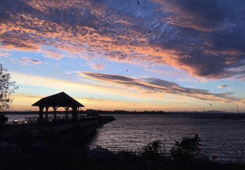 A serene waterfront scene at sunset, featuring a gazebo, colorful clouds, and birds in the sky.