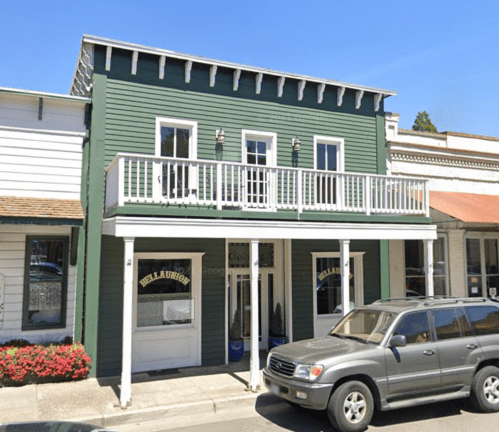 A green two-story building with a balcony, surrounded by flowers and parked cars on a sunny day.