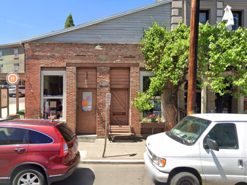 A brick building with a wooden door, surrounded by trees and parked cars on a sunny day.
