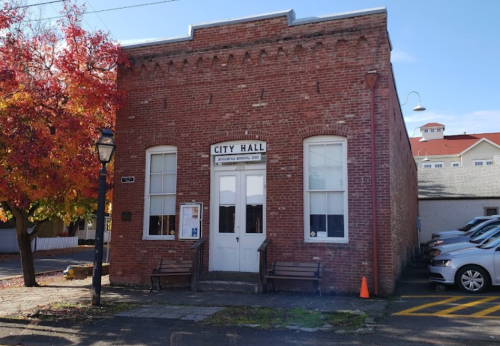 Brick building labeled "City Hall" with benches outside, surrounded by autumn trees and parked cars.