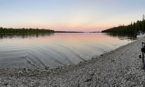 A serene lakeside view at sunset, with calm waters and a rocky shore lined by trees.