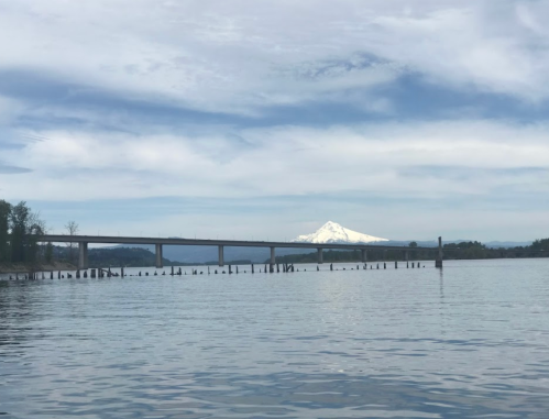 A serene river scene with a bridge and a snow-capped mountain in the background under a cloudy sky.