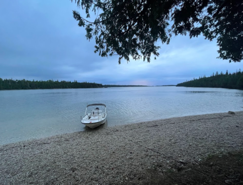 A calm lake scene at dusk with a boat on a pebbly shore and trees lining the water's edge.