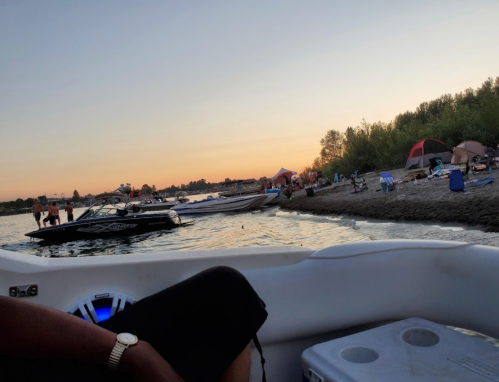 A sunset view of a river with boats and people on the shore, enjoying a beach day.