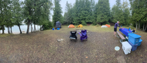 A campsite with tents, a picnic table, and chairs near a lake, surrounded by trees on a cloudy day.