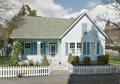 A light blue house with white trim, a picket fence, and a well-maintained yard under a partly cloudy sky.
