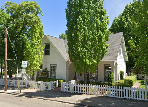 A white house with a gray roof, surrounded by tall green trees and a white picket fence on a sunny day.