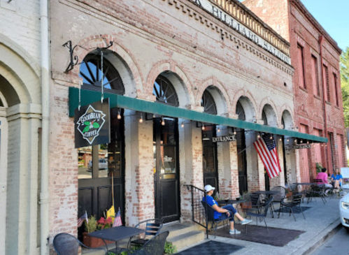 A charming coffee shop with arched windows, outdoor seating, and American flags, set in a historic brick building.