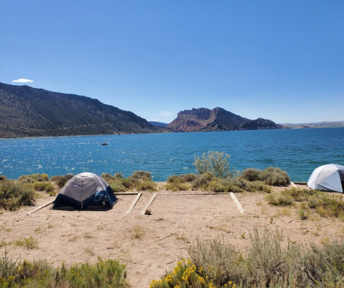 Two tents set up by a lake, surrounded by shrubs and mountains under a clear blue sky.
