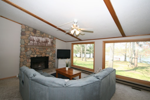 Cozy living room with a stone fireplace, large windows, and a sectional sofa, overlooking a scenic outdoor view.