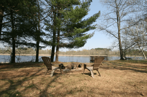 Two chairs facing a fire pit by a calm lake, surrounded by trees on a sunny day.