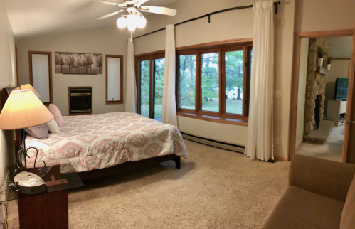 Cozy bedroom with a large window, a bed with a patterned quilt, a lamp, and a sofa, surrounded by nature.