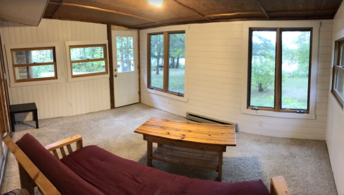 Cozy interior of a cabin with a wooden table, futon, and large windows overlooking a natural landscape.