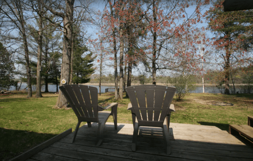 Two wooden chairs on a deck overlook a serene lake surrounded by trees and greenery.