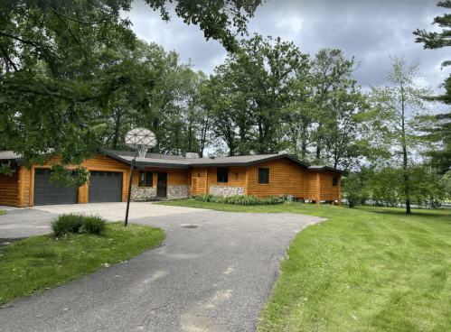 A modern wooden house surrounded by trees, featuring a driveway and a basketball hoop in the front yard.