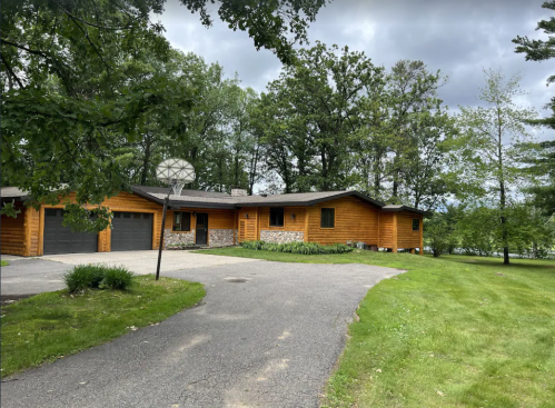 A modern wooden house with a stone facade, surrounded by trees, featuring a basketball hoop and a paved driveway.