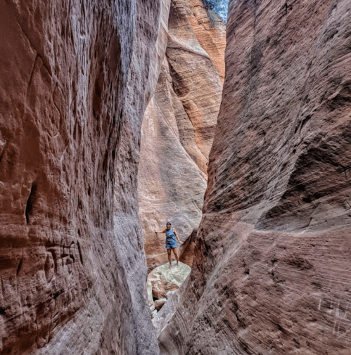 A person stands on a rock in a narrow canyon, surrounded by tall, textured sandstone walls.