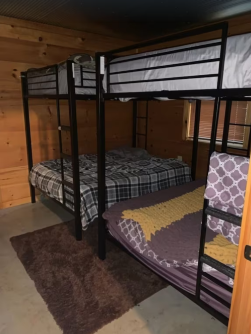 A cozy room with two sets of black metal bunk beds and a rug on a concrete floor. Wooden walls add warmth.