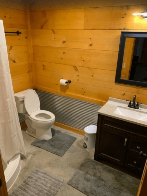 A cozy bathroom with wooden walls, a toilet, sink, shower curtain, and gray rugs on a concrete floor.