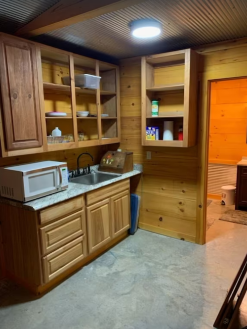 A rustic kitchen with wooden cabinets, a microwave, and a sink, leading to a bathroom in the background.