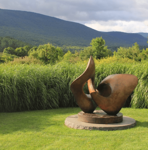 A bronze sculpture with flowing shapes sits on a stone base, surrounded by lush green grass and distant mountains.