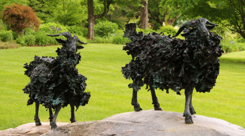 Two bronze goat sculptures covered in textured foliage, standing on a rock in a lush green garden.
