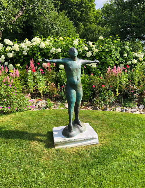 A bronze sculpture of a standing figure with outstretched arms, surrounded by vibrant flowers and greenery.