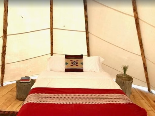 Cozy tent interior with a bed, decorative blanket, and wooden stools, creating a warm, inviting atmosphere.