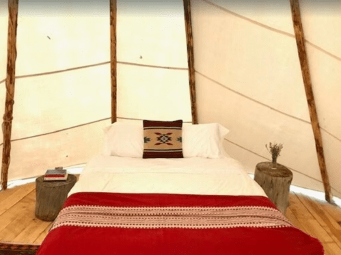 Cozy tent interior with a bed, decorative blanket, and wooden stools, creating a warm, inviting atmosphere.