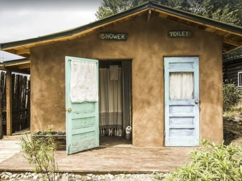 A rustic building with two doors labeled "Shower" and "Toilet," featuring a wooden deck and greenery around it.