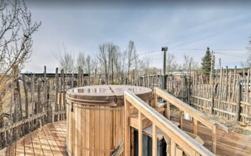 A wooden hot tub on a deck surrounded by a rustic fence and trees under a clear sky.